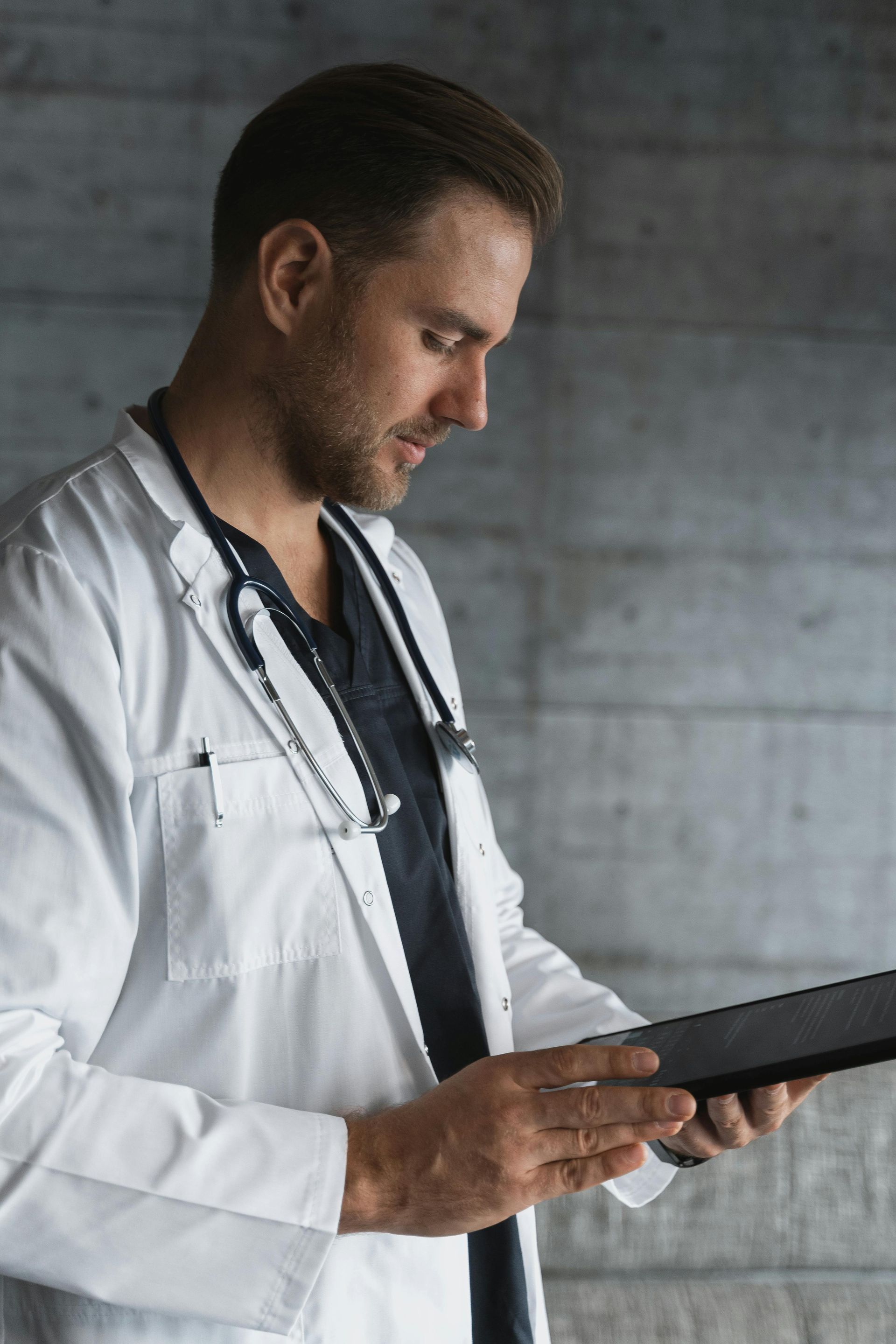 Doctor in white coat, stethoscope, looking down at tablet in front of concrete wall.