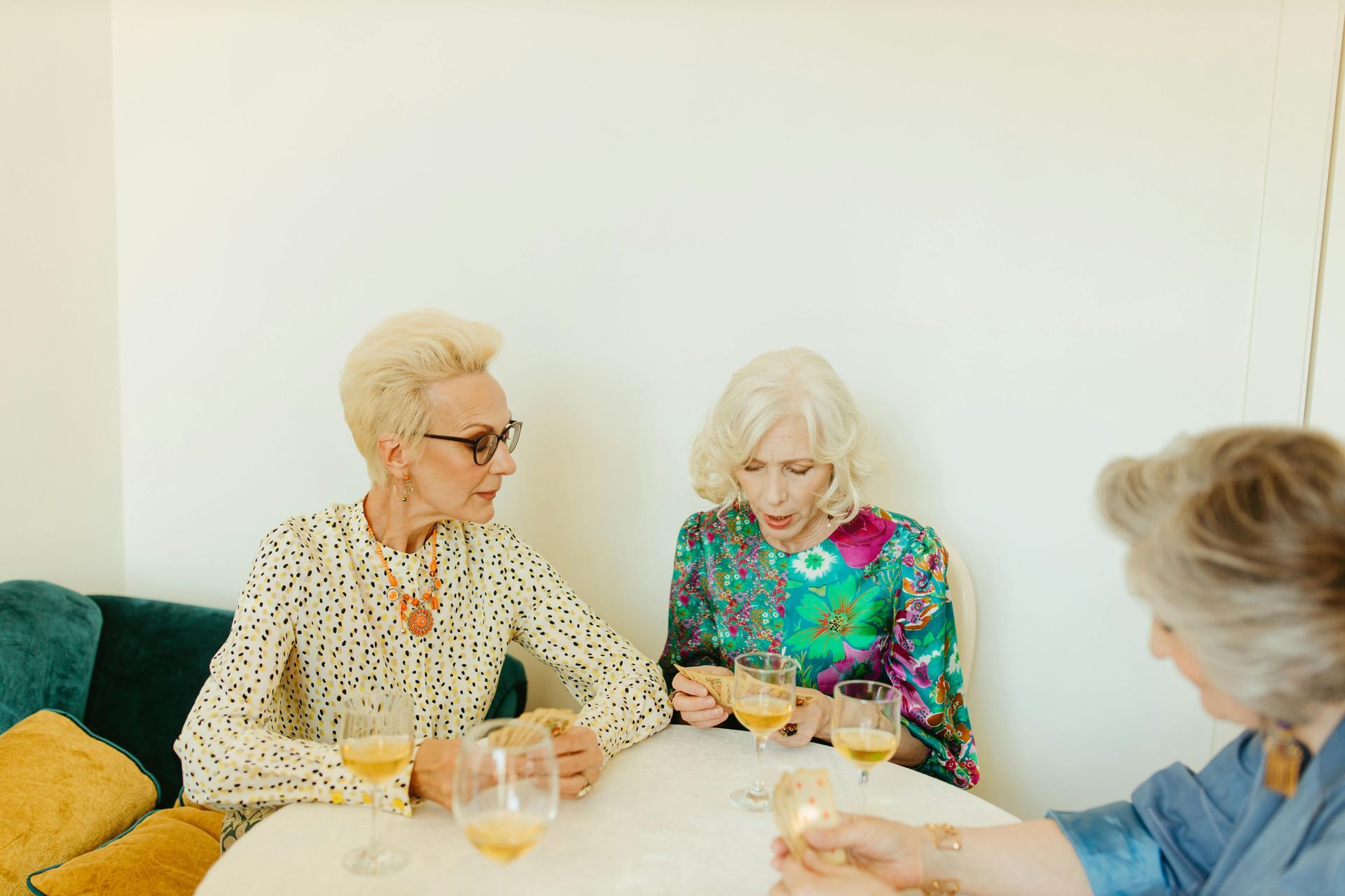Three stylish older women at a table, drinking champagne, one looking surprised, light and bright setting.