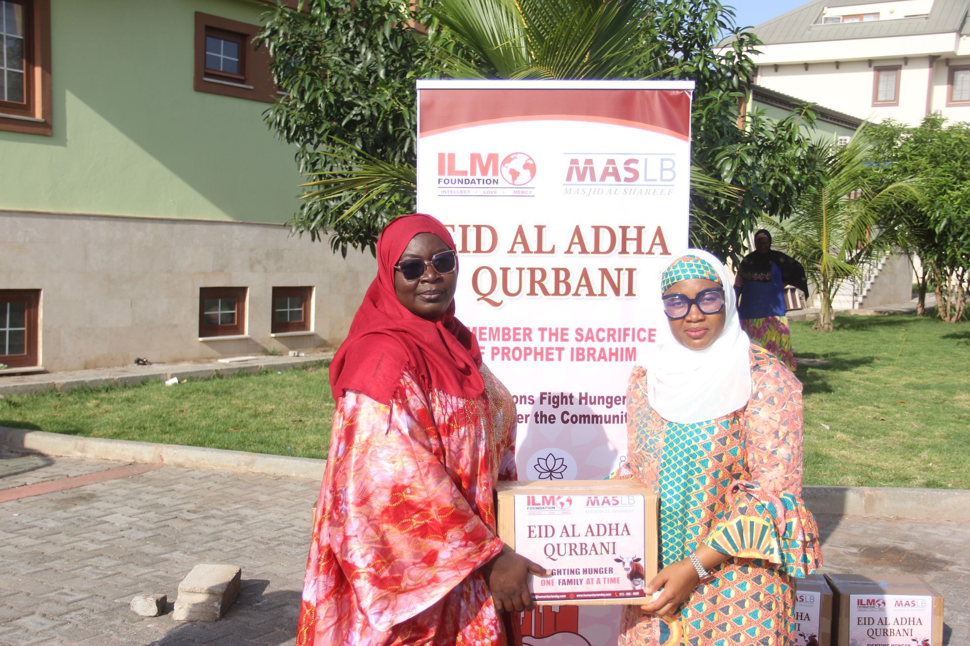 Two women standing in front of a sign that says id al adha qurbani