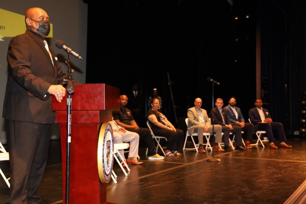 A man wearing a mask is standing at a podium in front of a group of people sitting in chairs.