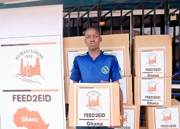 A young man holding a box of food donations in front of stacked boxes labeled 