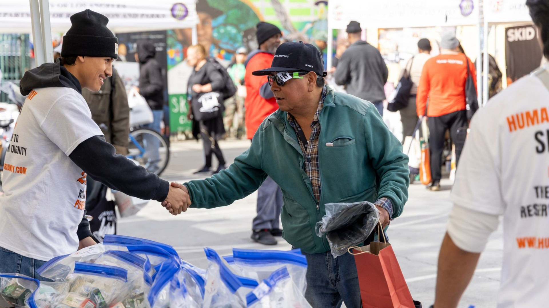 Two men at a vendor stand, looking at rolled fabrics. A tent is behind them, with a building mural visible.