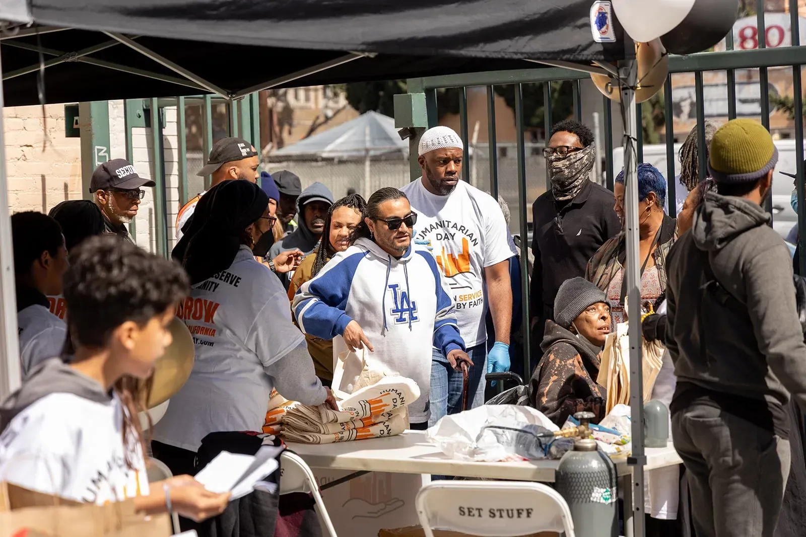 People gather outdoors at a table, some wearing white shirts, possibly an event.