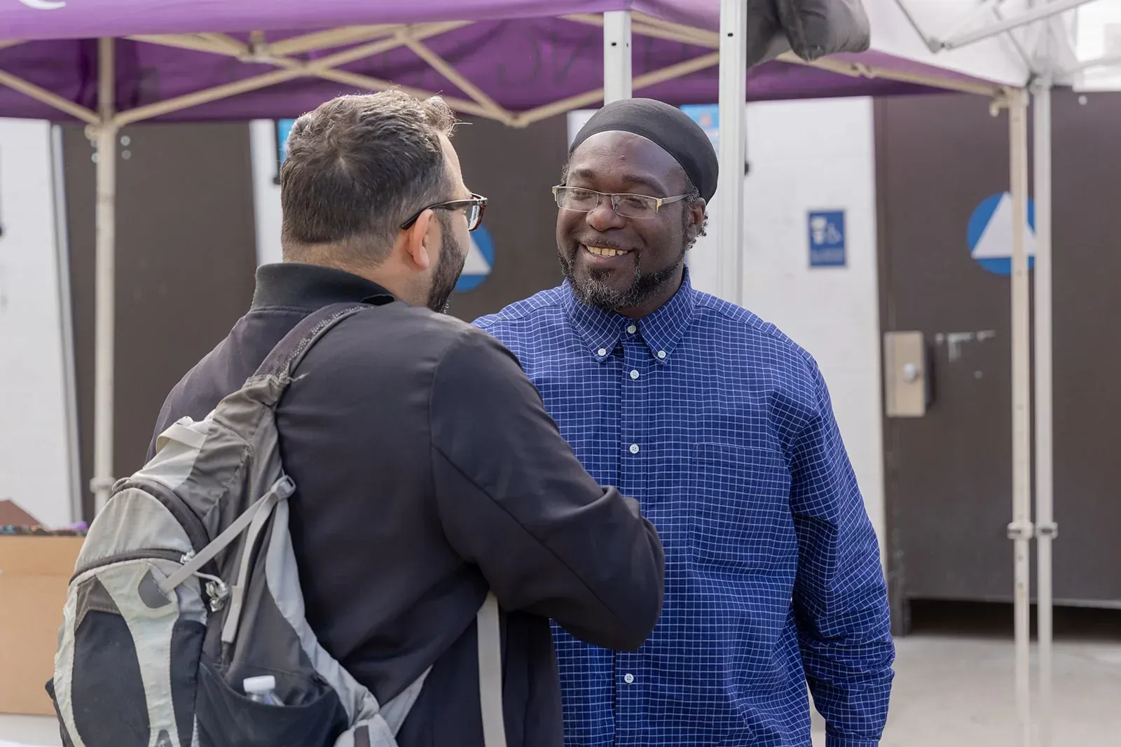 Two men greeting each other, one with glasses and a backpack, the other smiling wearing a blue shirt and a hat.