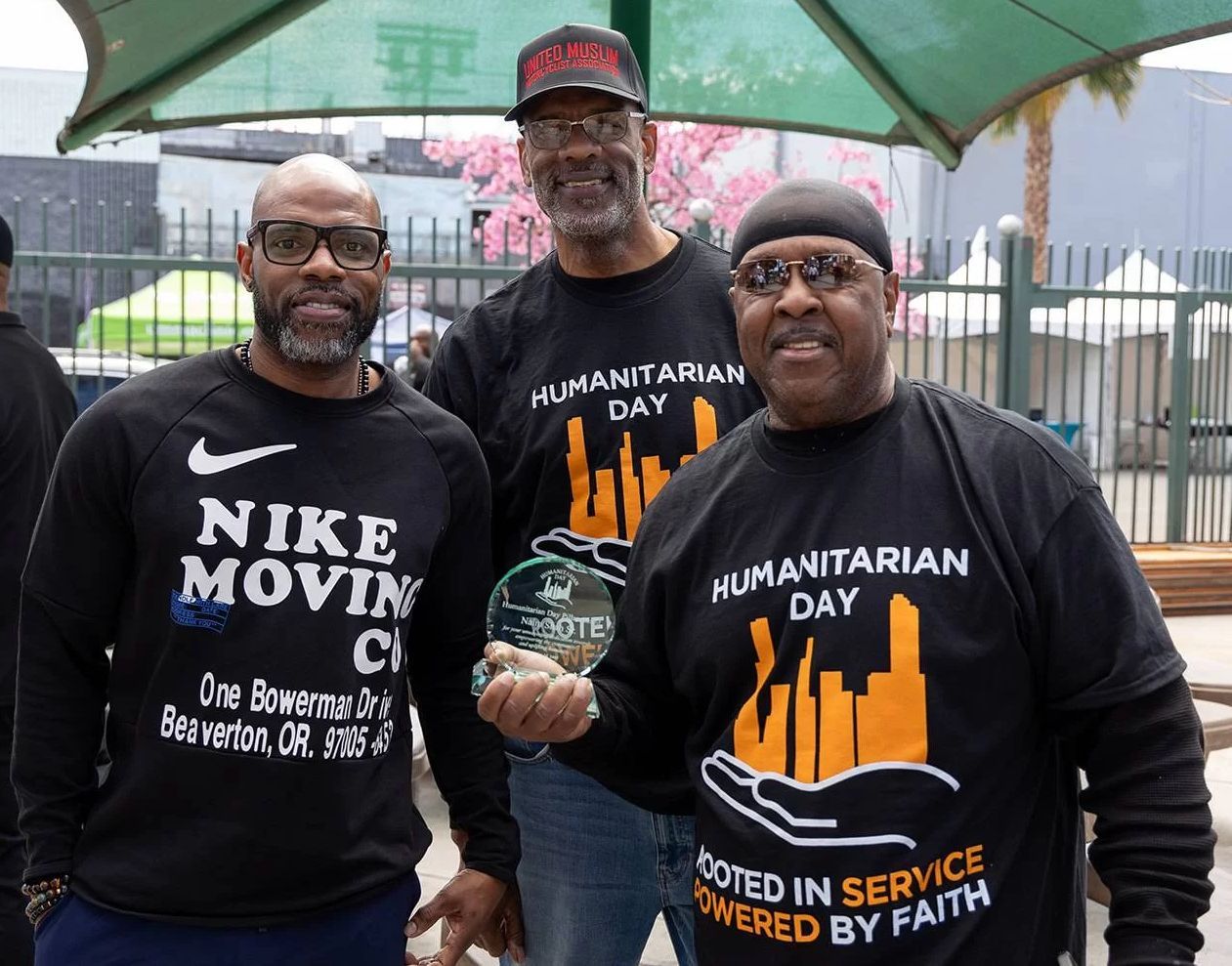 Three Black men at Humanitarian Day event, smiling. One holds a trophy; they wear shirts with logos.
