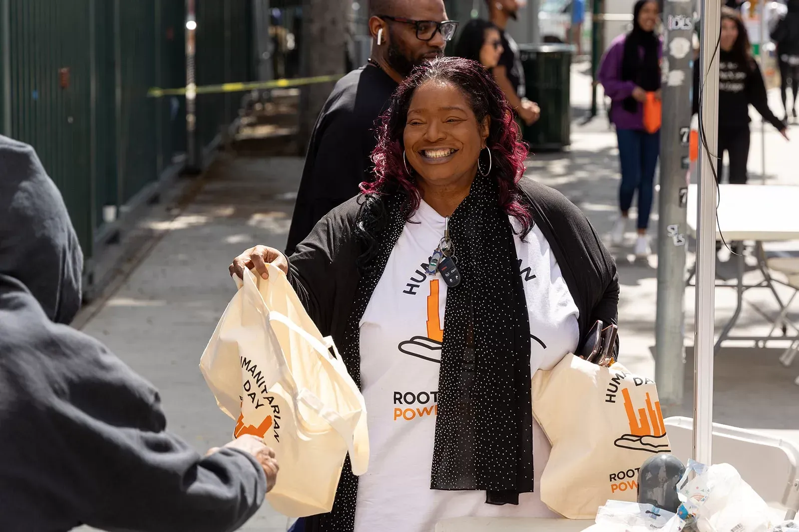 Woman with a black scarf and a white shirt smiles while handing out bags. Street scene.