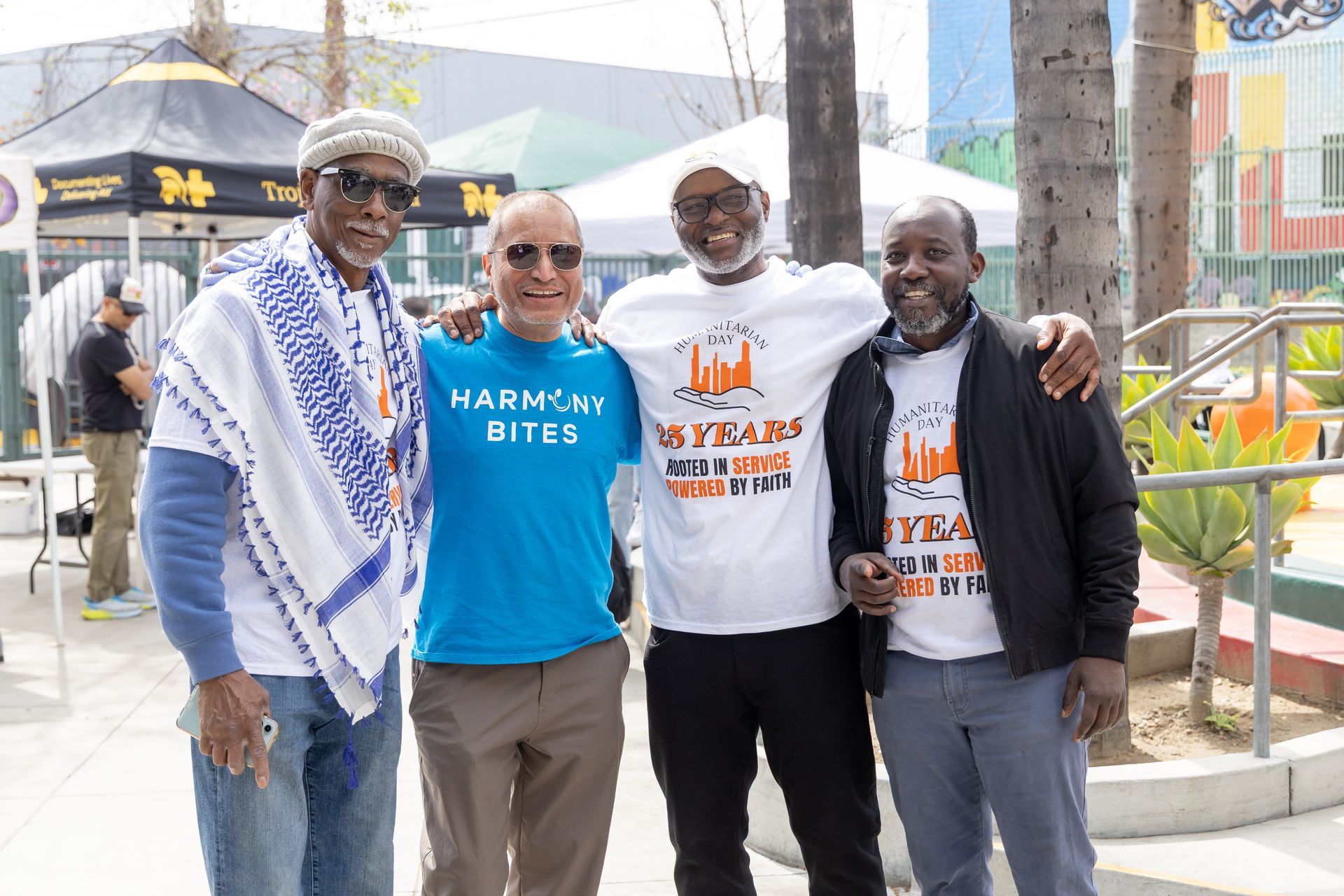 Four men pose outdoors, smiling. One wears a blue shirt, the others white shirts with text. Background: tents, buildings.