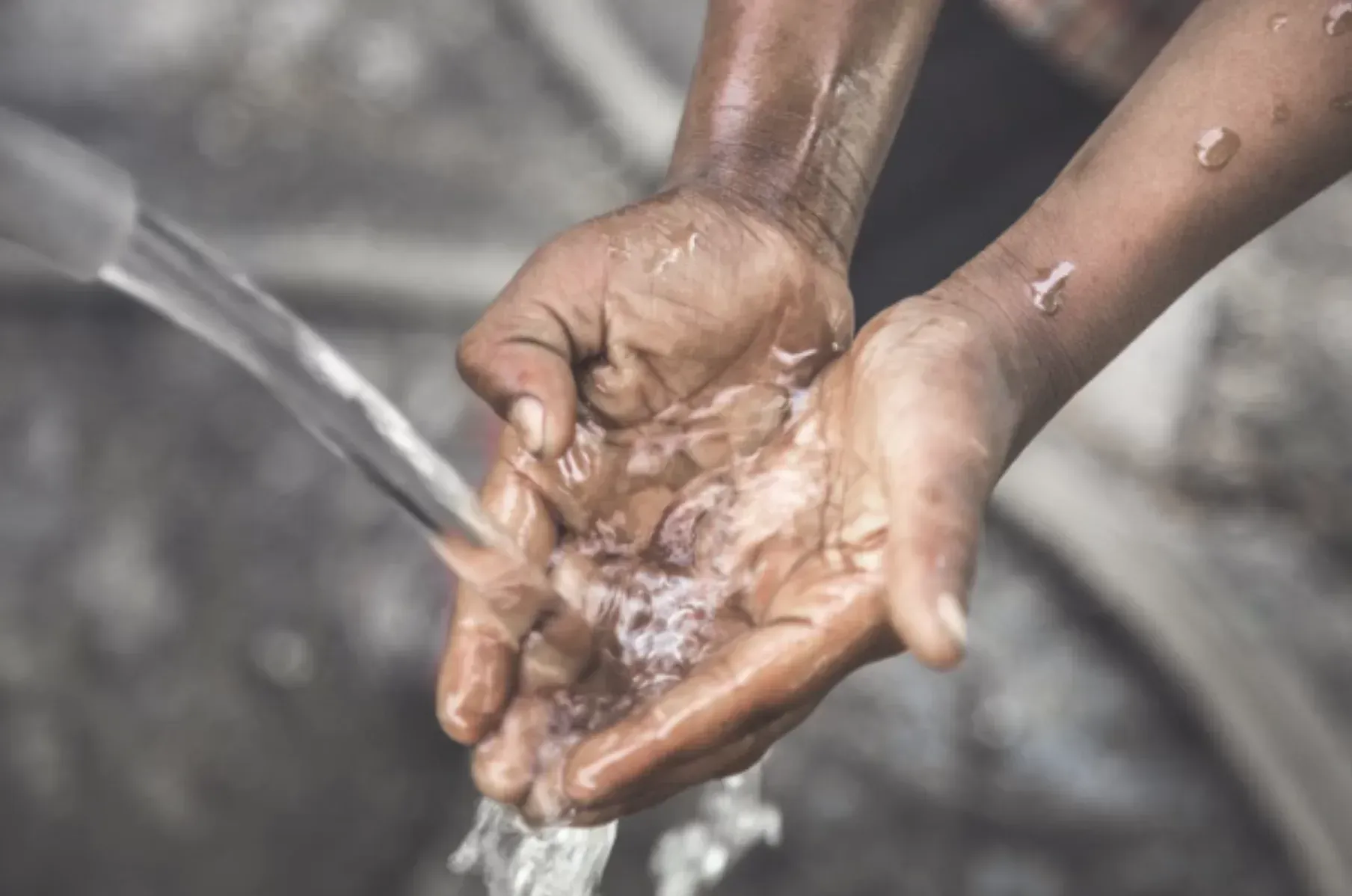 Dark-skinned hands cupped under running water. Bubbles and droplets visible, suggesting washing outdoors.