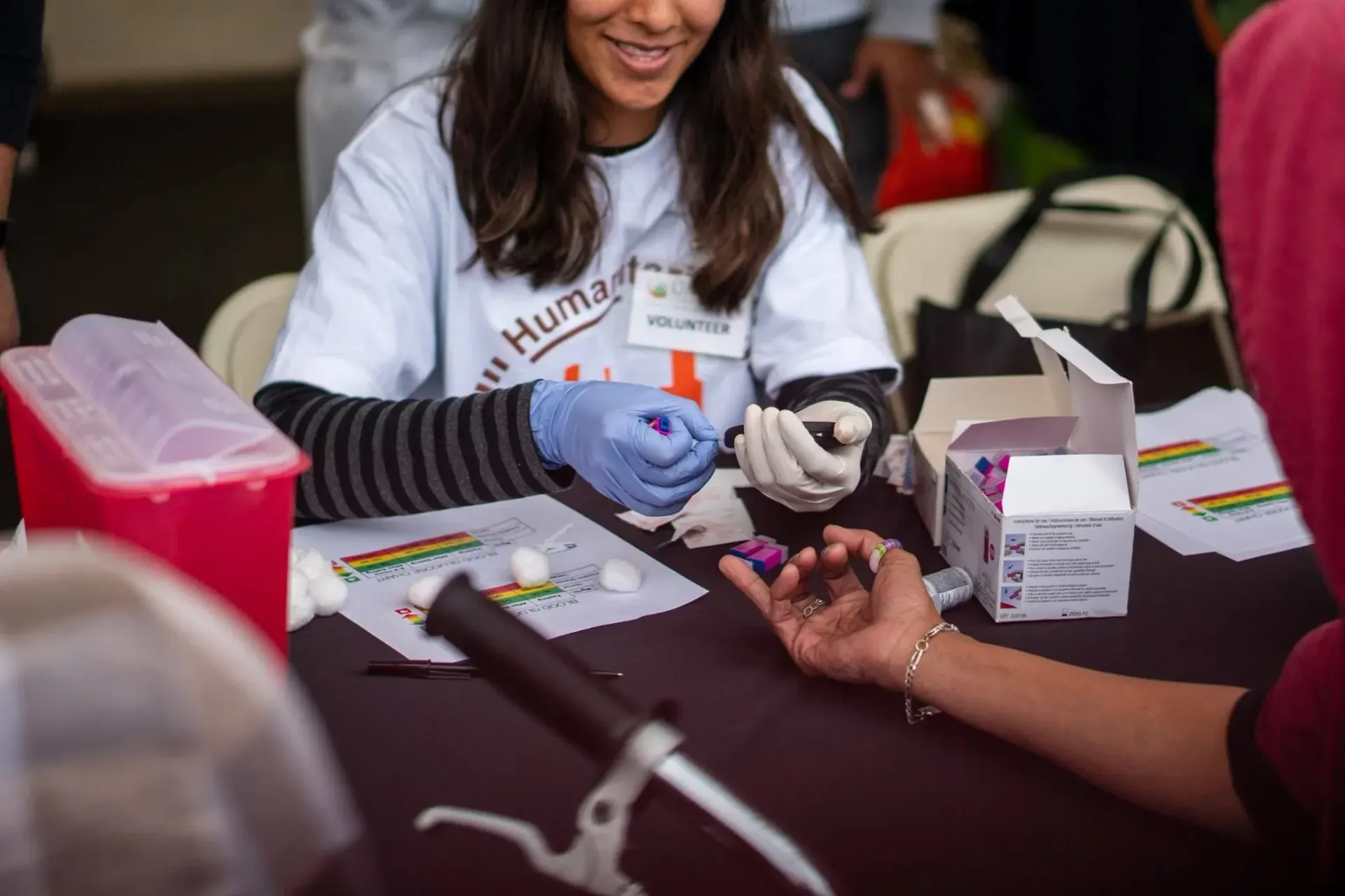 Person taking a finger prick blood sample at a table. Woman wearing a white shirt and gloves is administering the test.