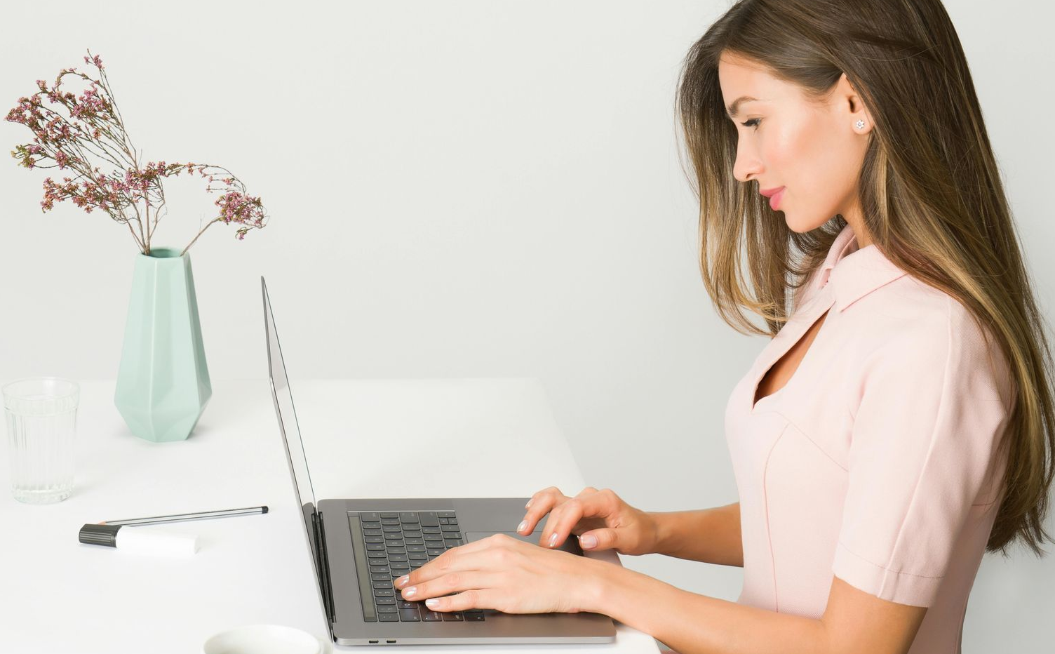 Woman working on laptop at a white desk, wearing a light pink shirt, with flowers and a vase to the left.
