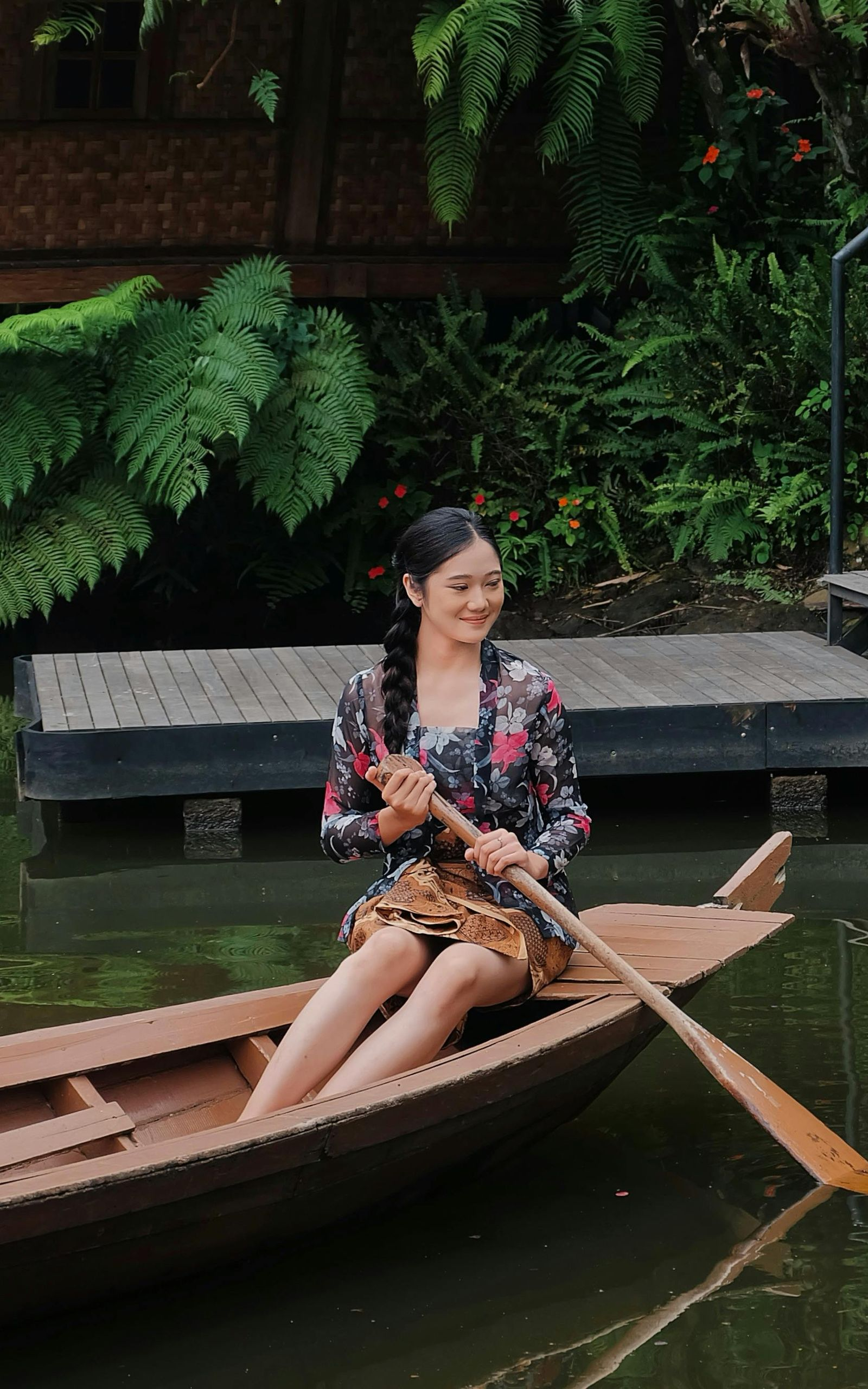 Woman in a wooden boat, smiling, paddling on a waterway. Lush greenery and wooden structures in the background.