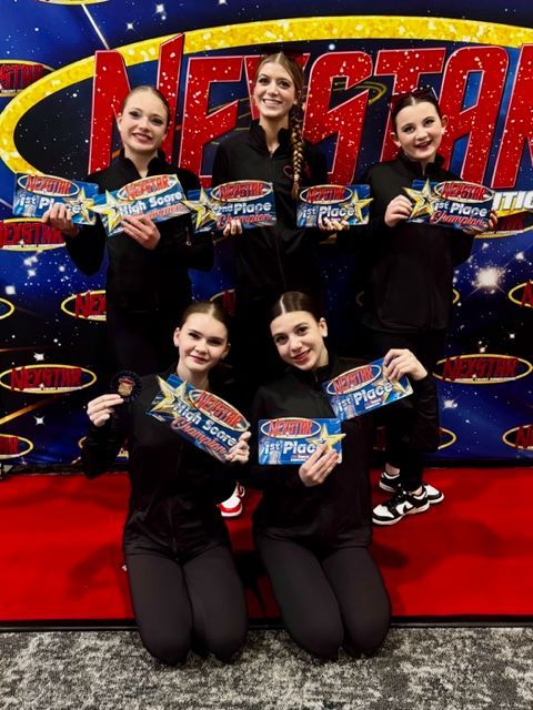 Five young female dancers in black outfits hold awards, posing on a red carpet.