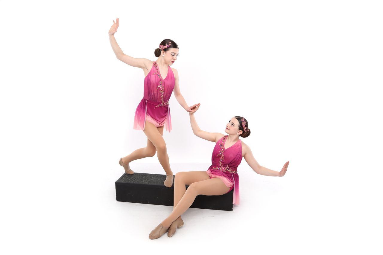 Two dancers in pink costumes. One steps on a black block, reaching toward the other sitting on the ground.