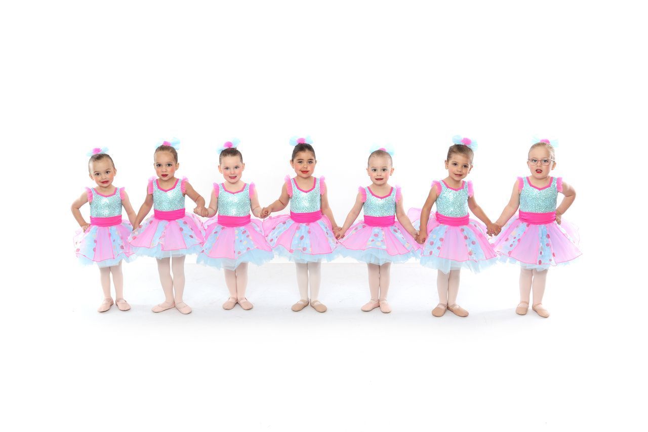 Seven young girls in colorful tutus and matching headbands holding hands, posing against a white background.