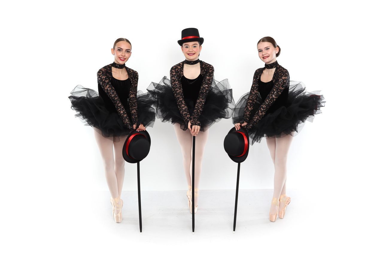 Three ballet dancers in black tutus, holding canes and top hats, smiling against a white background.