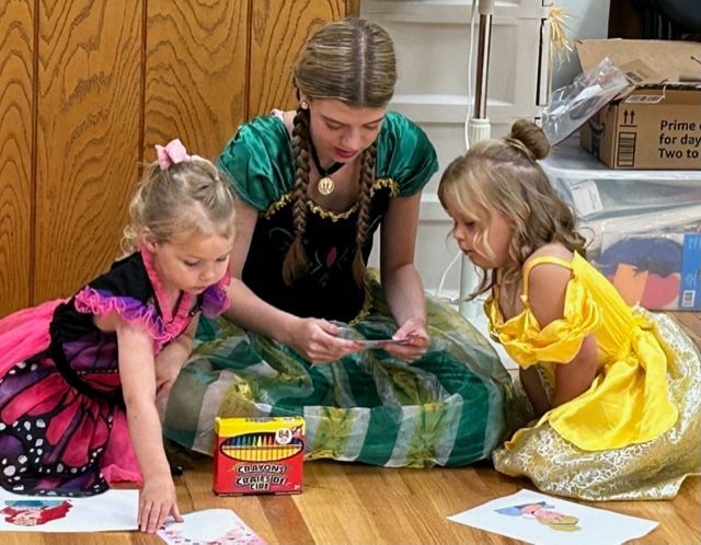 Woman and two children in princess costumes drawing on the floor with crayons.