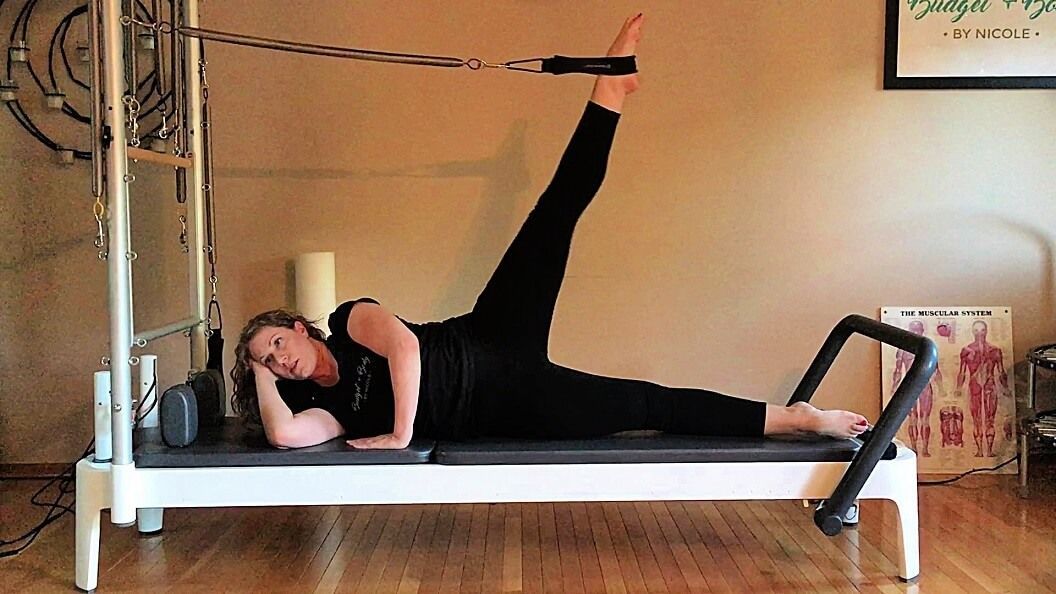 Woman on Pilates reformer, leg extended, attached to strap. Beige wall background.