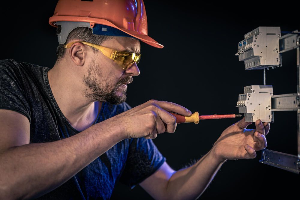 Electrician in hard hat and safety glasses, using screwdriver on electrical panel.
