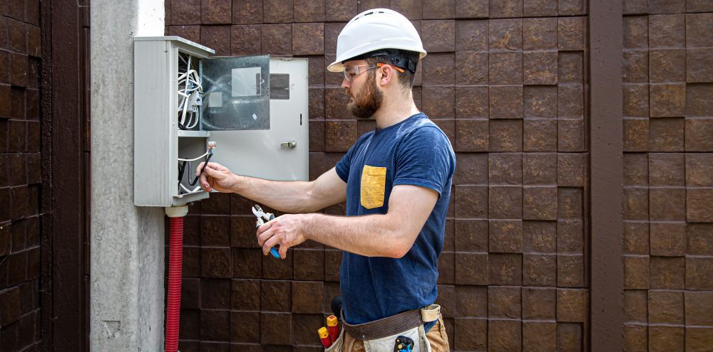 Electrician working on electrical panel, wearing a hard hat, outside.