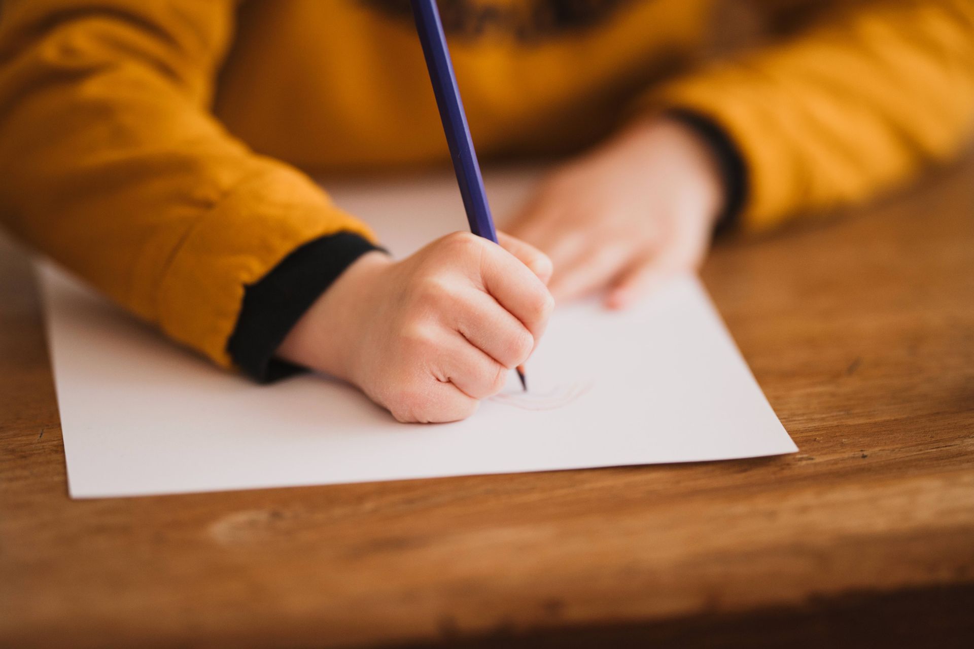 Gros plan sur la main d'un enfant tenant un crayon violet, écrivant sur du papier blanc. Chemise jaune, table en bois.