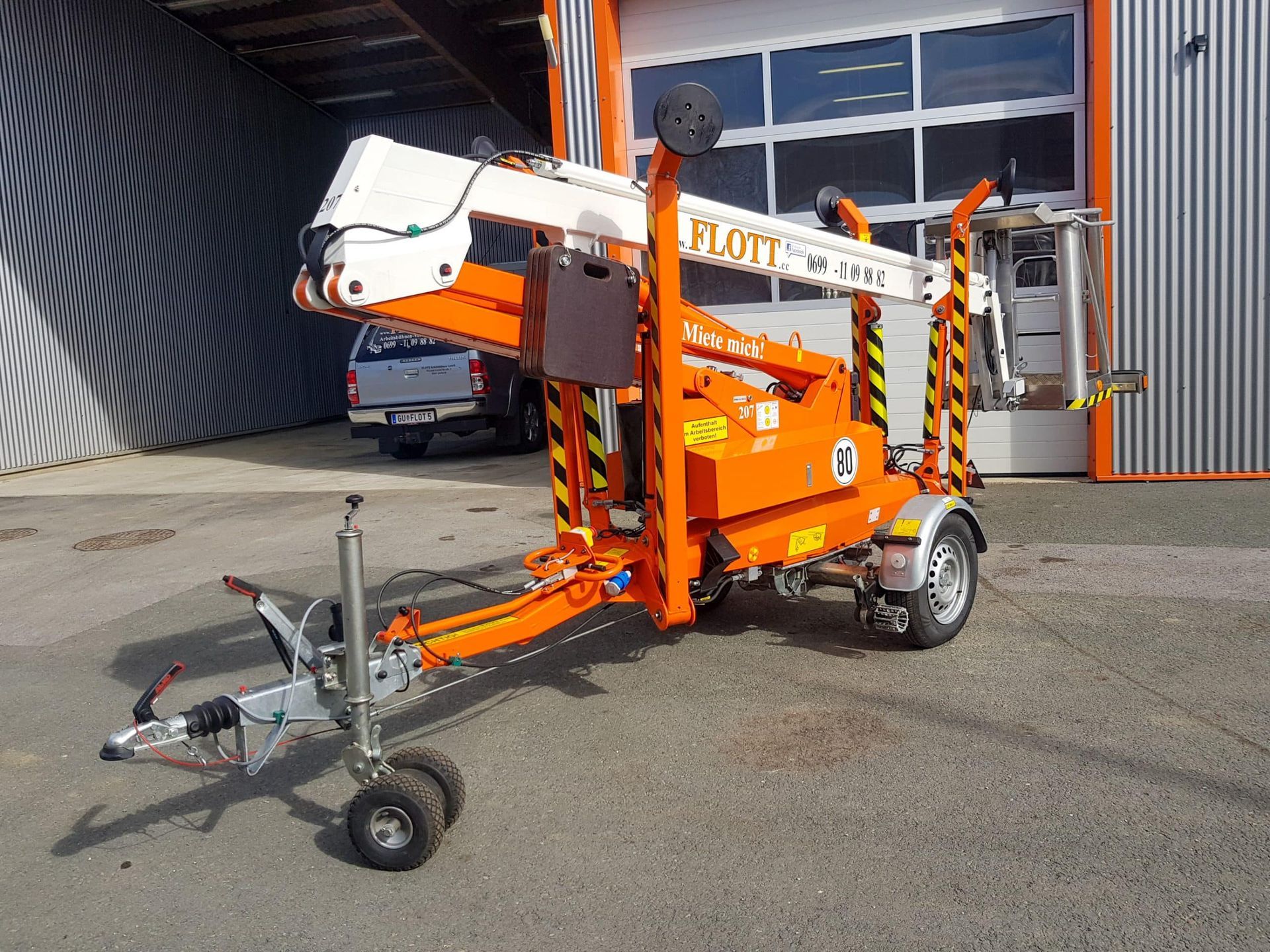 Eine orange-weiße Flott-Hubarbeitsbühne auf einem Anhänger, der vor einem Gebäude geparkt ist.