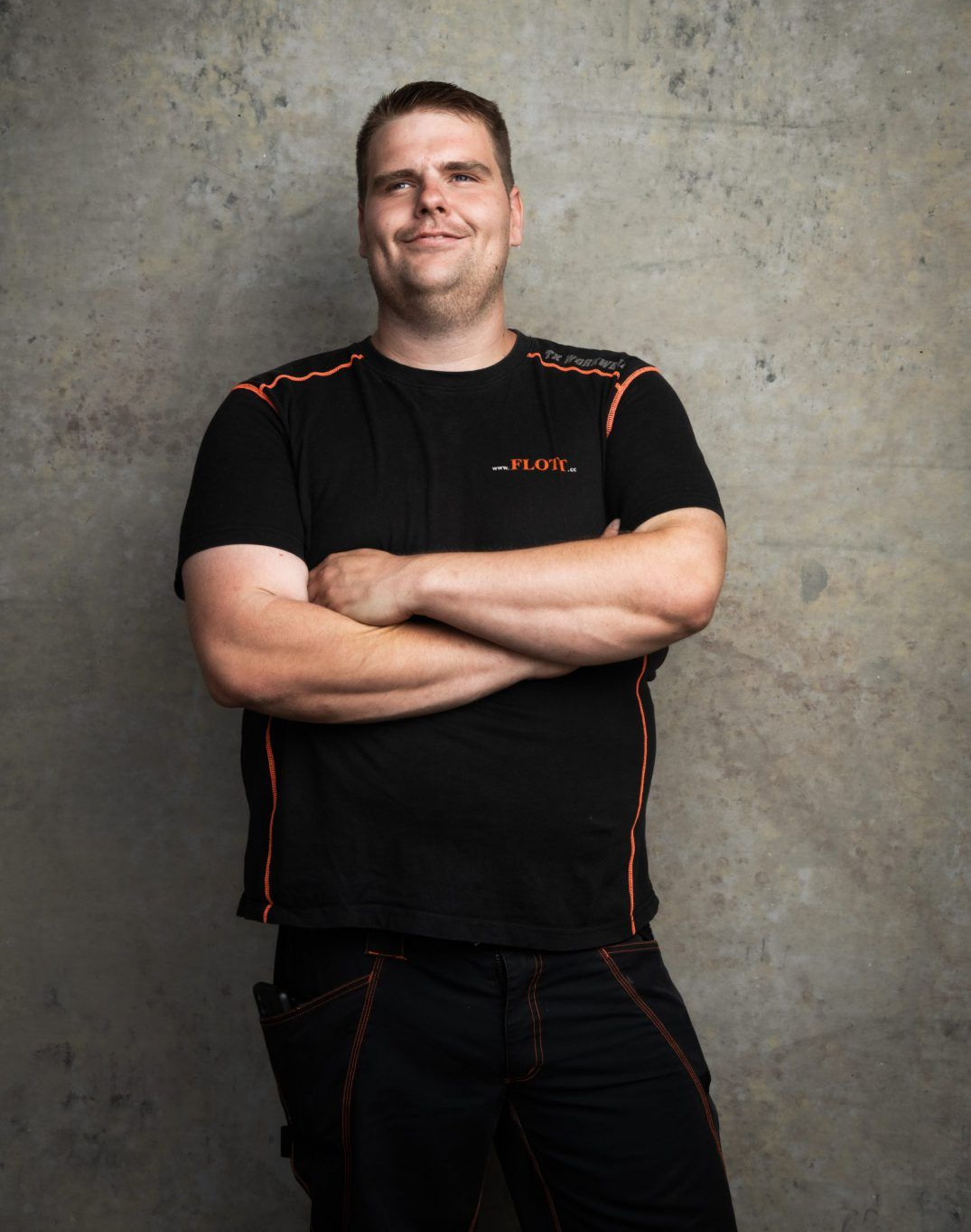 Mann in schwarzer Arbeitskleidung mit verschränkten Armen, der an einer Betonwand lehnt.