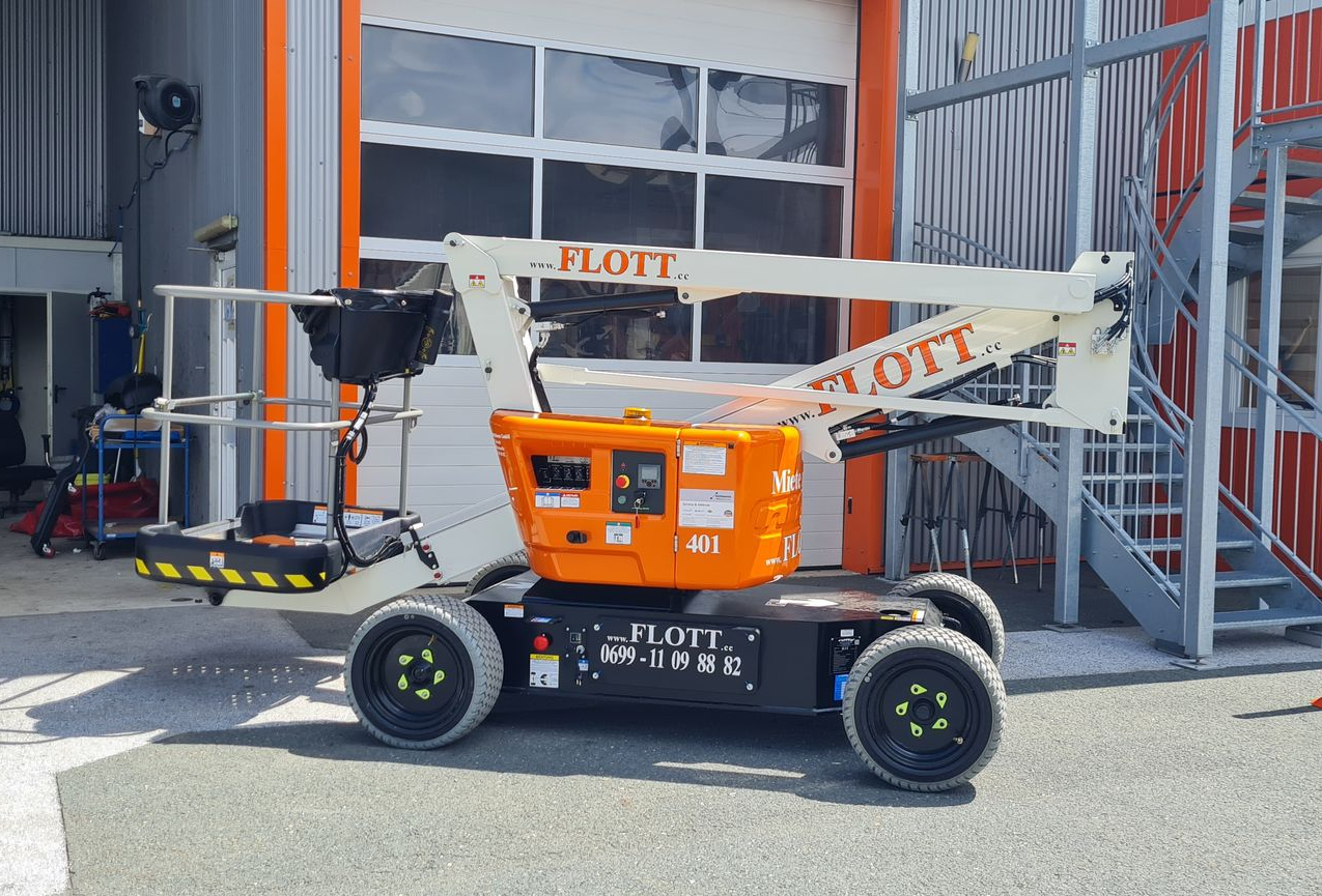 Eine orange-weiße JLG-Arbeitsbühne mit schwarzen Reifen steht vor einem Gebäude mit orangefarbener Tür.