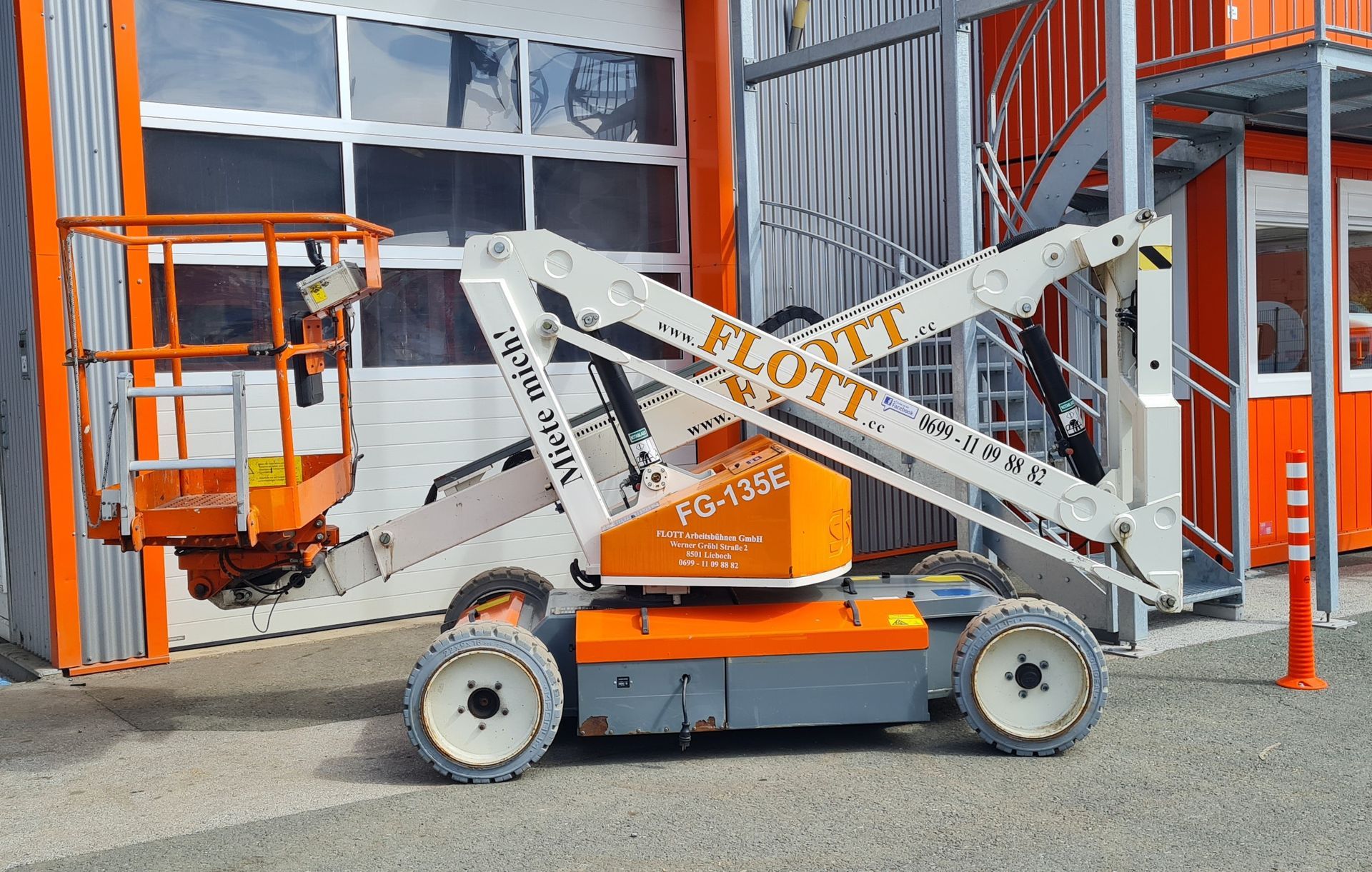 Eine orange-weiße Gelenkteleskoparbeitsbühne steht vor einem Gebäude.