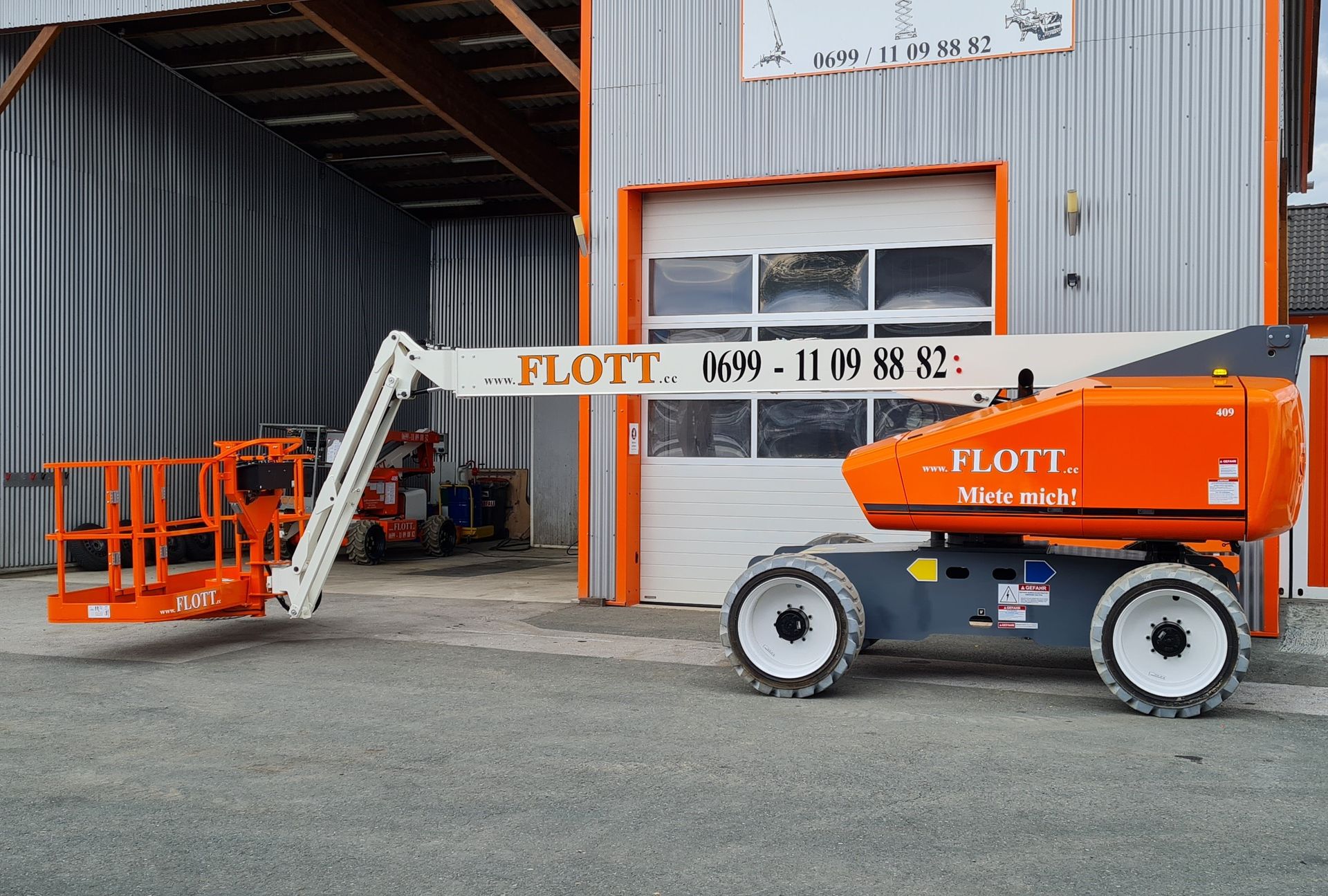 Eine orange-weiße Gelenkteleskoparbeitsbühne steht vor einem Metallgebäude mit Garagentor.