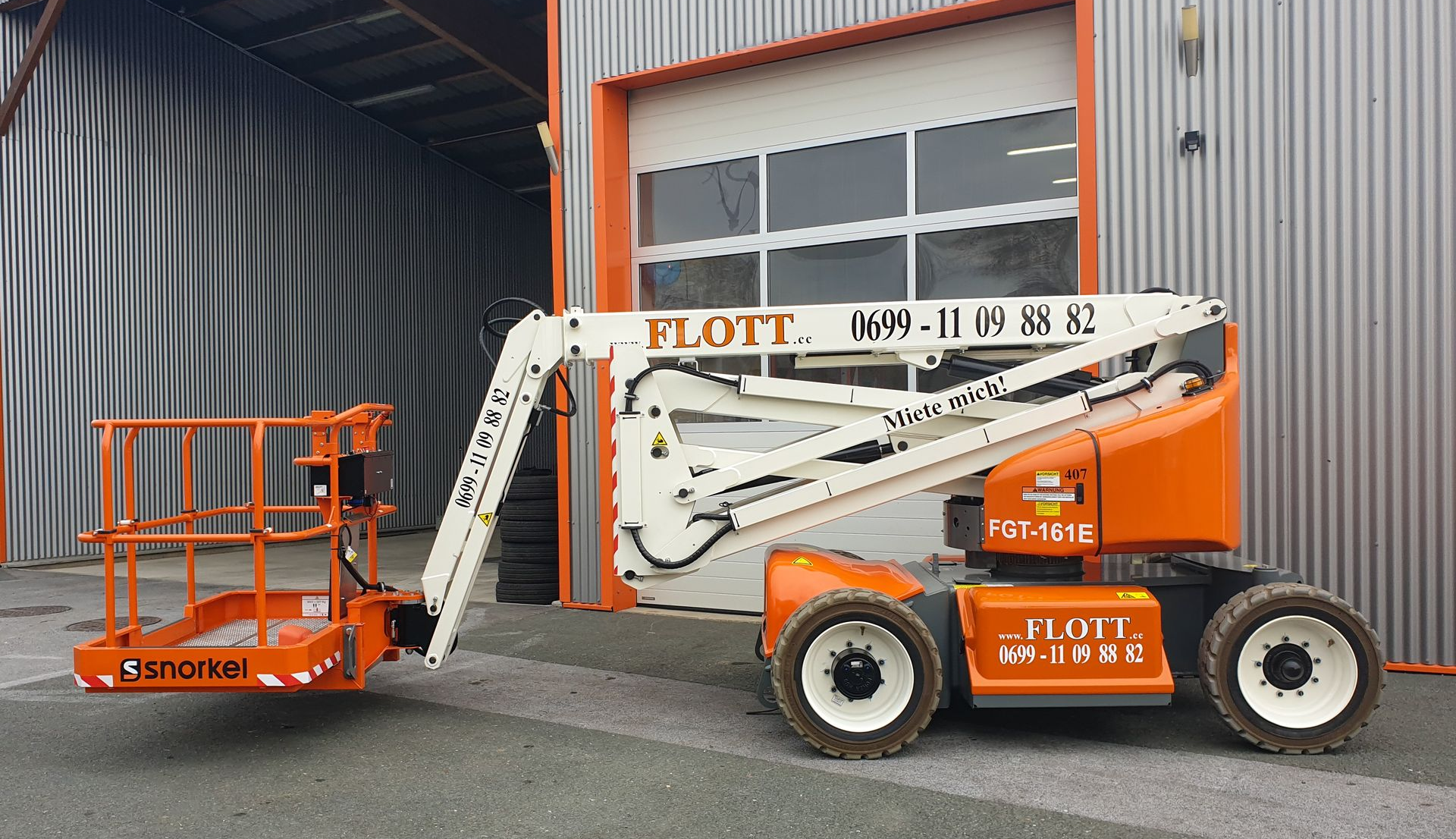 Eine orange-weiße FLOTT-Arbeitsbühne ist in der Nähe eines Gebäudes geparkt.