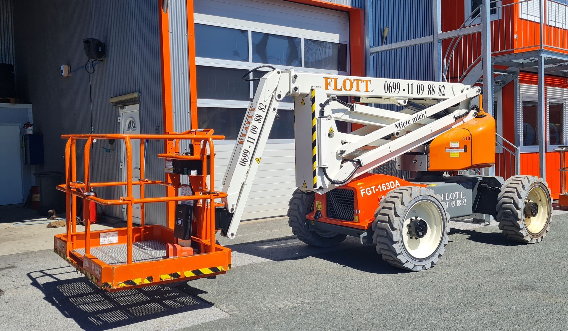 Orange-weiße Hubarbeitsbühne vor einem Gebäude; Hubarm ausgefahren, Korb leer.