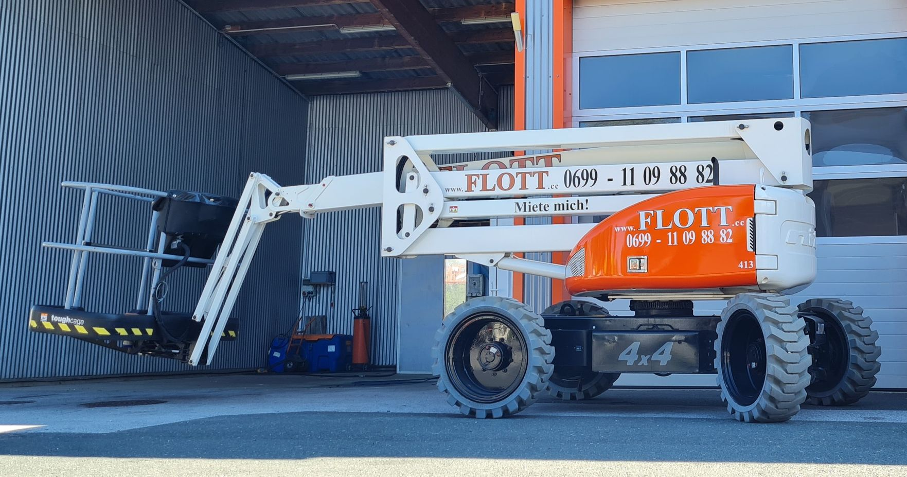 Orange und weiße JLG-Hubarbeitsbühne mit ausfahrbarem Arm und Plattform vor einer Wellblechkonstruktion.