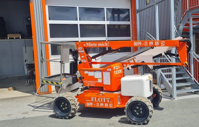 Eine orangefarbene FLOTT-Scherenhebebühne ist in der Nähe eines Gebäudes mit offenem Garagentor und Metalltreppe geparkt.