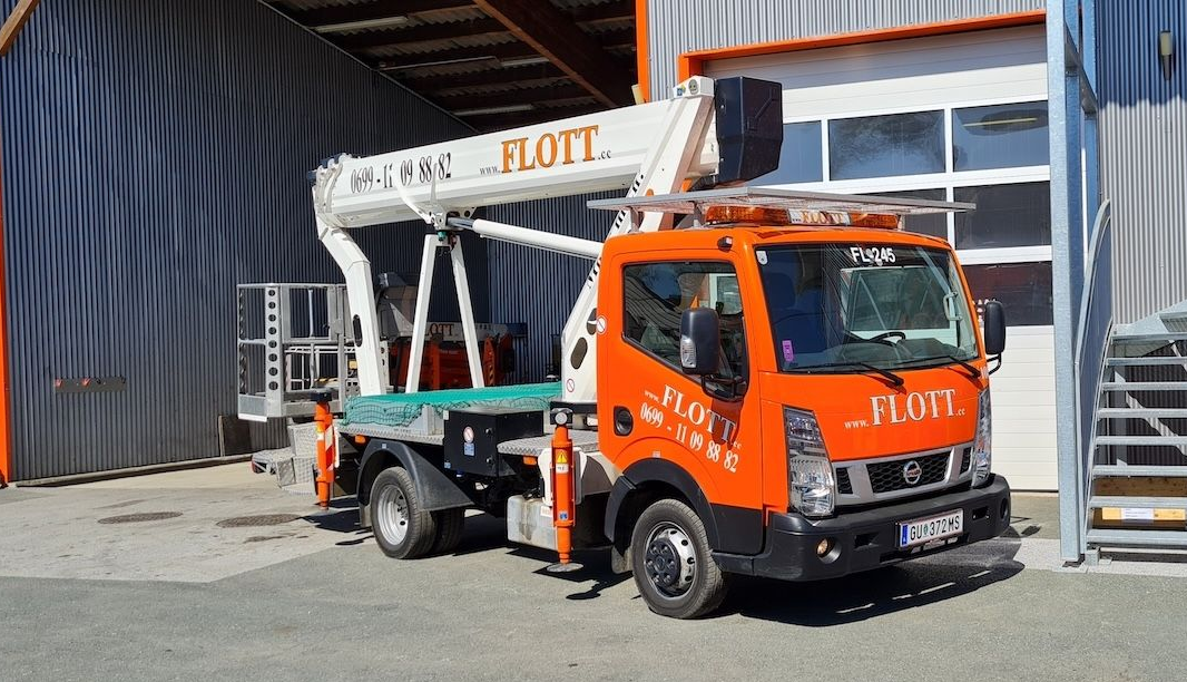 Ein orangefarbener Lkw mit ausgefahrenem Hubarm parkt vor einem Gebäude. Auf dem Lkw steht „FLOT“.