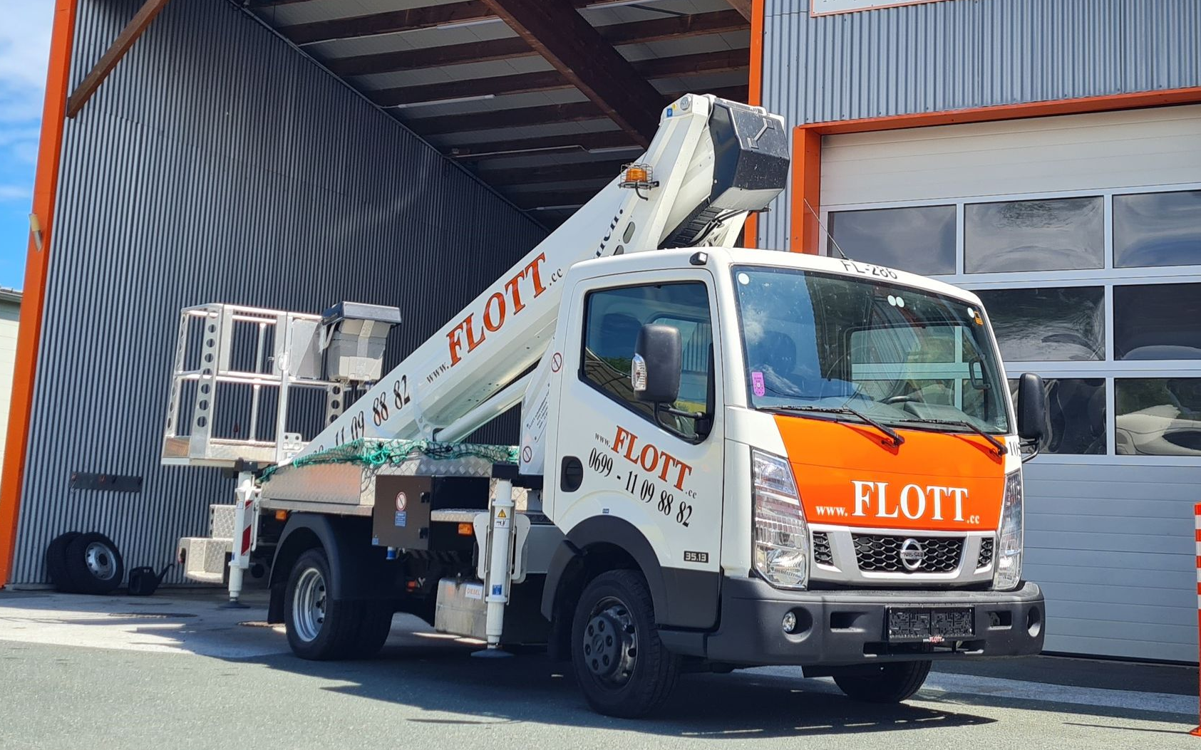 Weißer „FLOTT“-Lkw mit Hubarbeitsbühne, geparkt unter einem Unterstand. Orangefarbene Details an der Front.