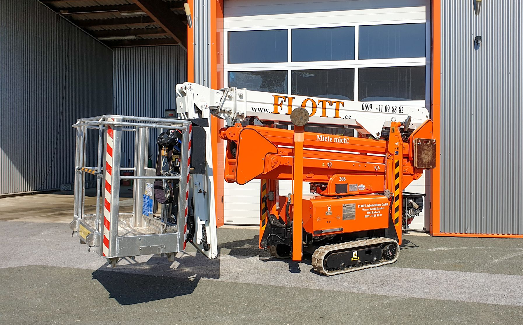 Orange-weiße Raupenarbeitsbühne mit angebautem Korb vor einem Gebäude.