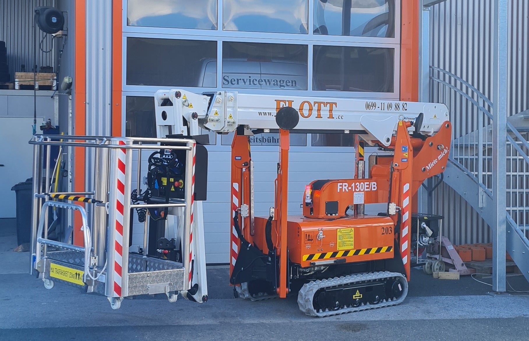 Ein orange-weißer Flott-Minilift mit Schienen, neben einer silbernen Arbeitsplattform, vor einer Garage.