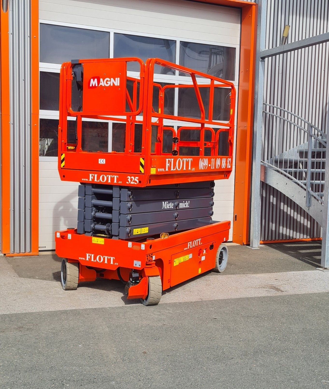 Eine orange-graue Scherenhebebühne lehnt an einem Gebäude mit geschlossenem Garagentor.