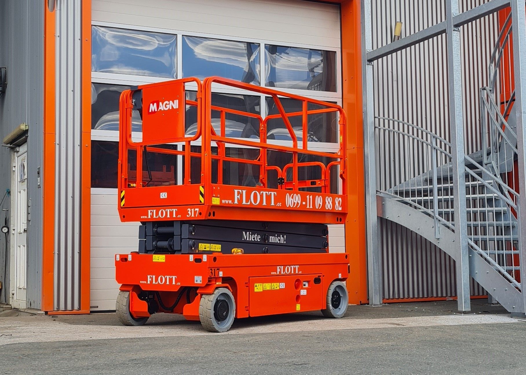 Eine orangefarbene Scherenhebebühne vor einem weißen Garagentor, neben einer Metalltreppe.