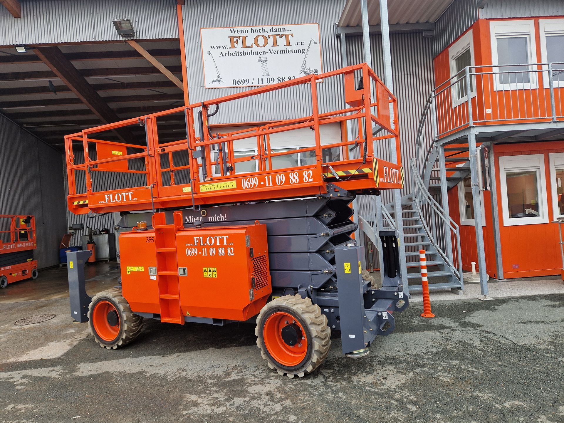 Eine orange-graue Scherenhebebühne auf einer Baustelle, in der Nähe eines Gebäudes und eines Treppenhauses.