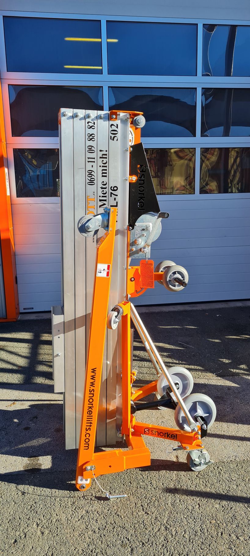 Ein orange-silberner, fahrbarer Aufzug vor einem Glasfenster.