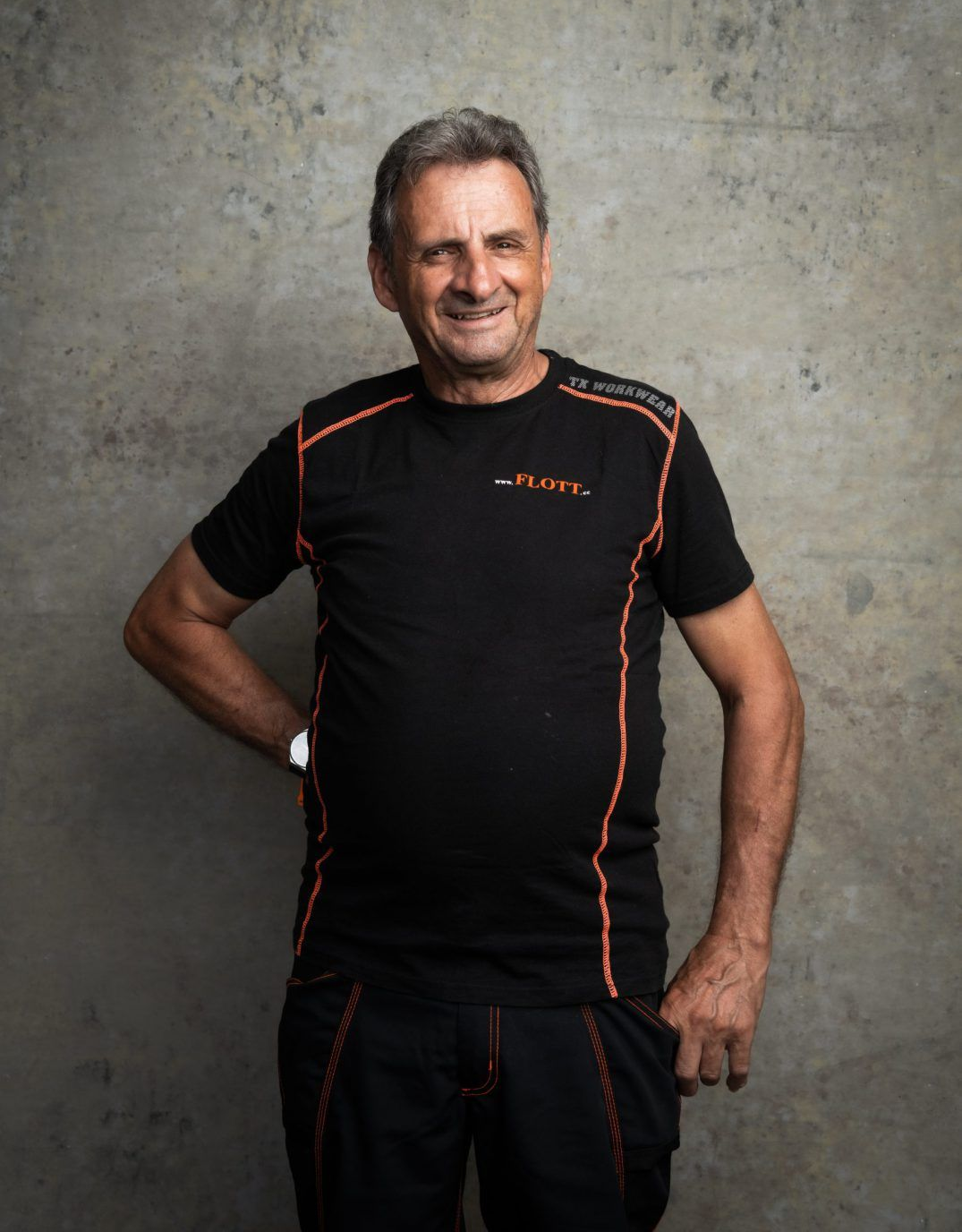 Mann in schwarzem Hemd mit orangefarbenem Saum, lächelnd, vor einer Betonwand. Hand in die Hüfte gestemmt, lässige Pose.