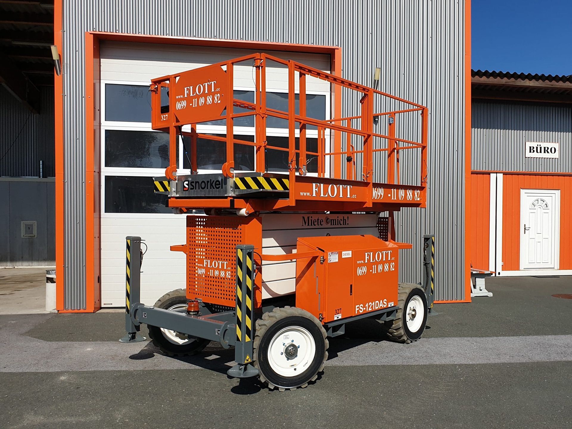 Eine orangefarbene Scherenhebebühne auf Rädern vor einem Metallgebäude.