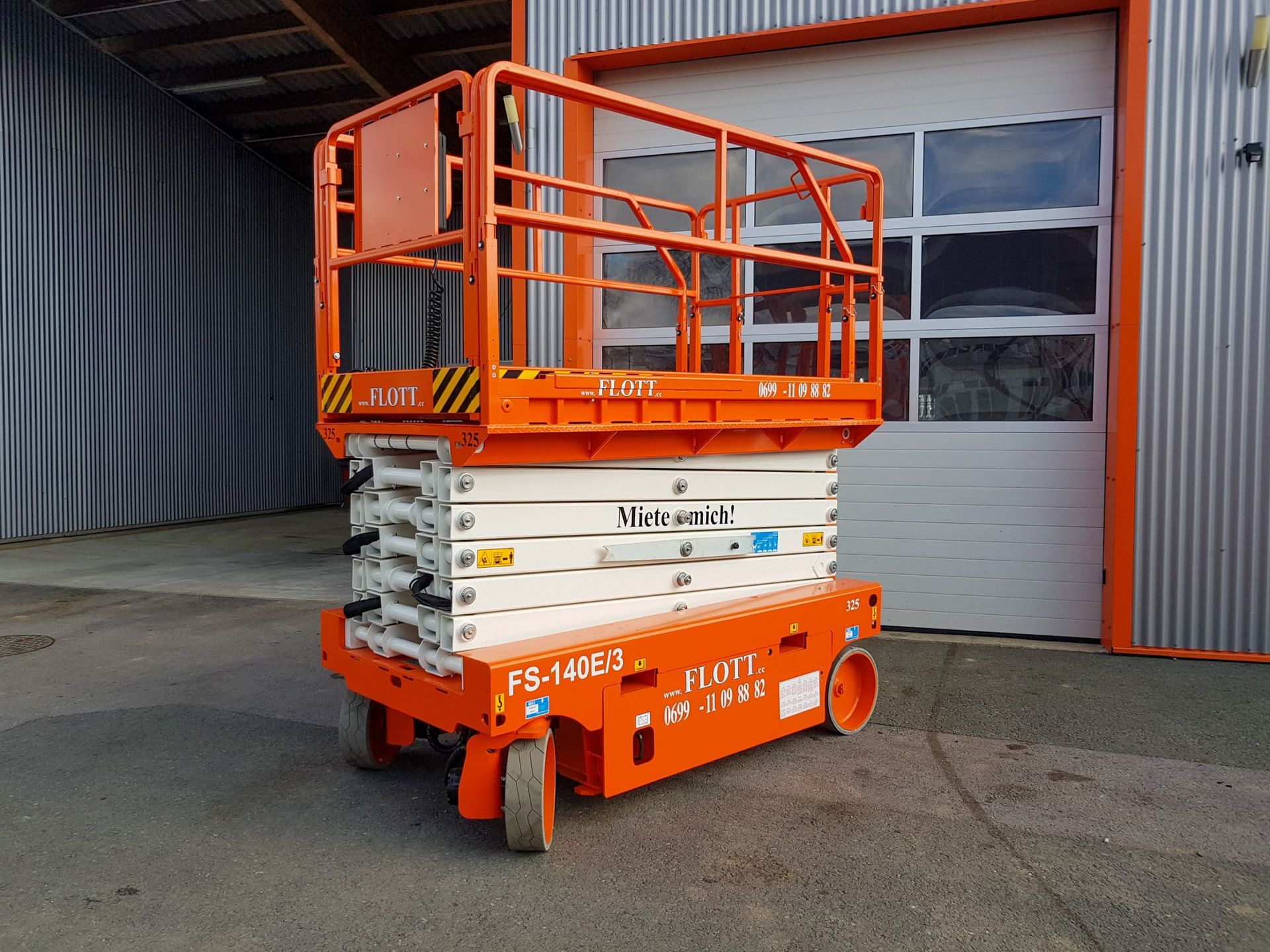 Eine orange-weiße Scherenhebebühne neben einem Wellblechgebäude mit Garagentor.