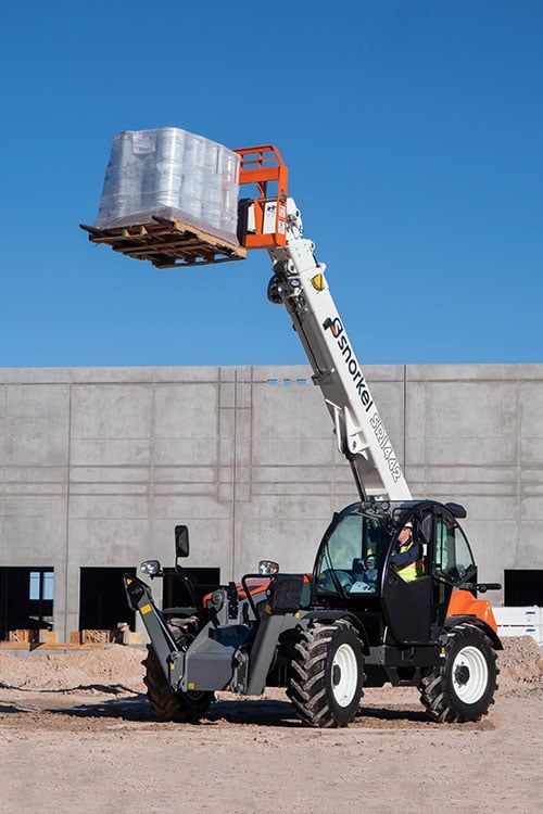 Ein Teleskoplader hebt eine Palette mit verpackten Baumaterialien an. Der Fahrer sitzt in der Kabine vor blauem Himmel.