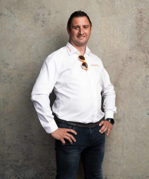 Mann in weißem Hemd und Jeans, Hände in die Hüften gestemmt, lächelt. Sonnenbrille auf dem Hemd.