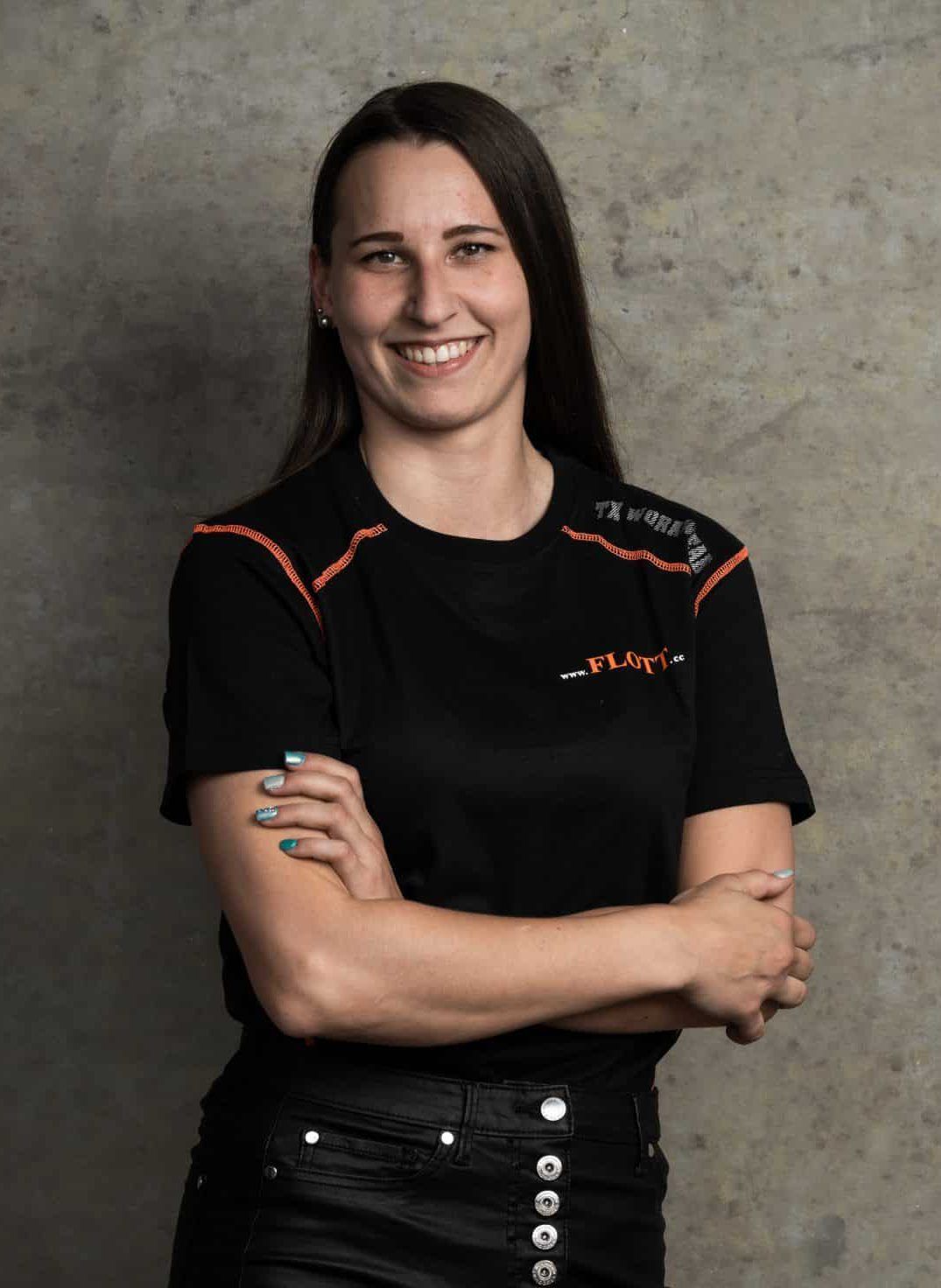 Eine lächelnde Frau mit verschränkten Armen, die ein schwarzes Hemd mit orangefarbenen Akzenten steht vor einer grauen Wand.