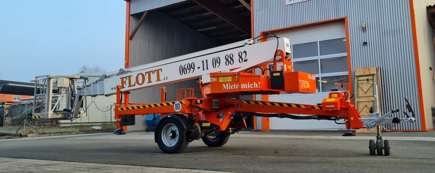Eine orangefarbene Flott-Hebebühne steht im Freien vor einem hellen Gebäude.