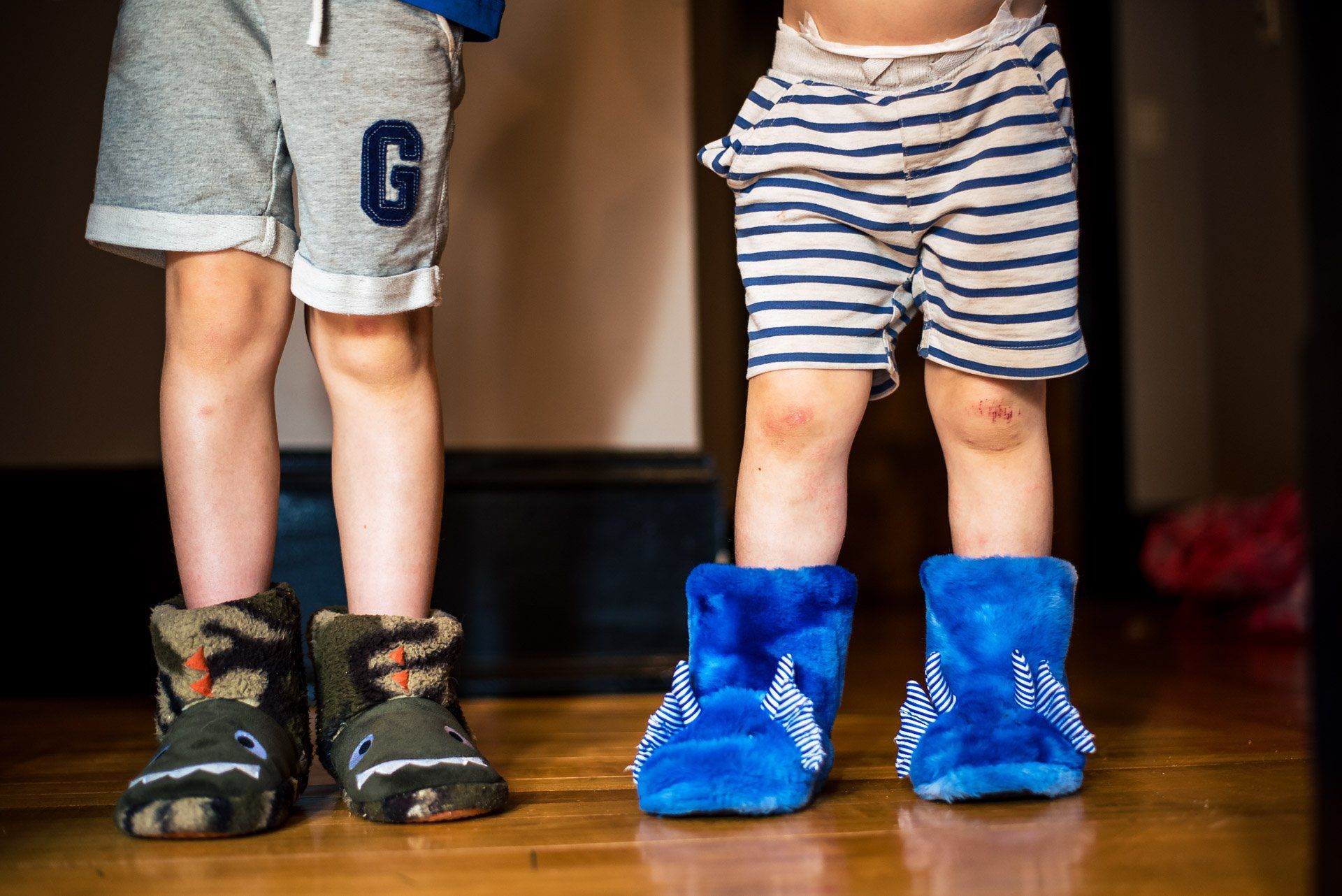 Two young boys wearing slippers and shorts are standing next to each other on a wooden floor.