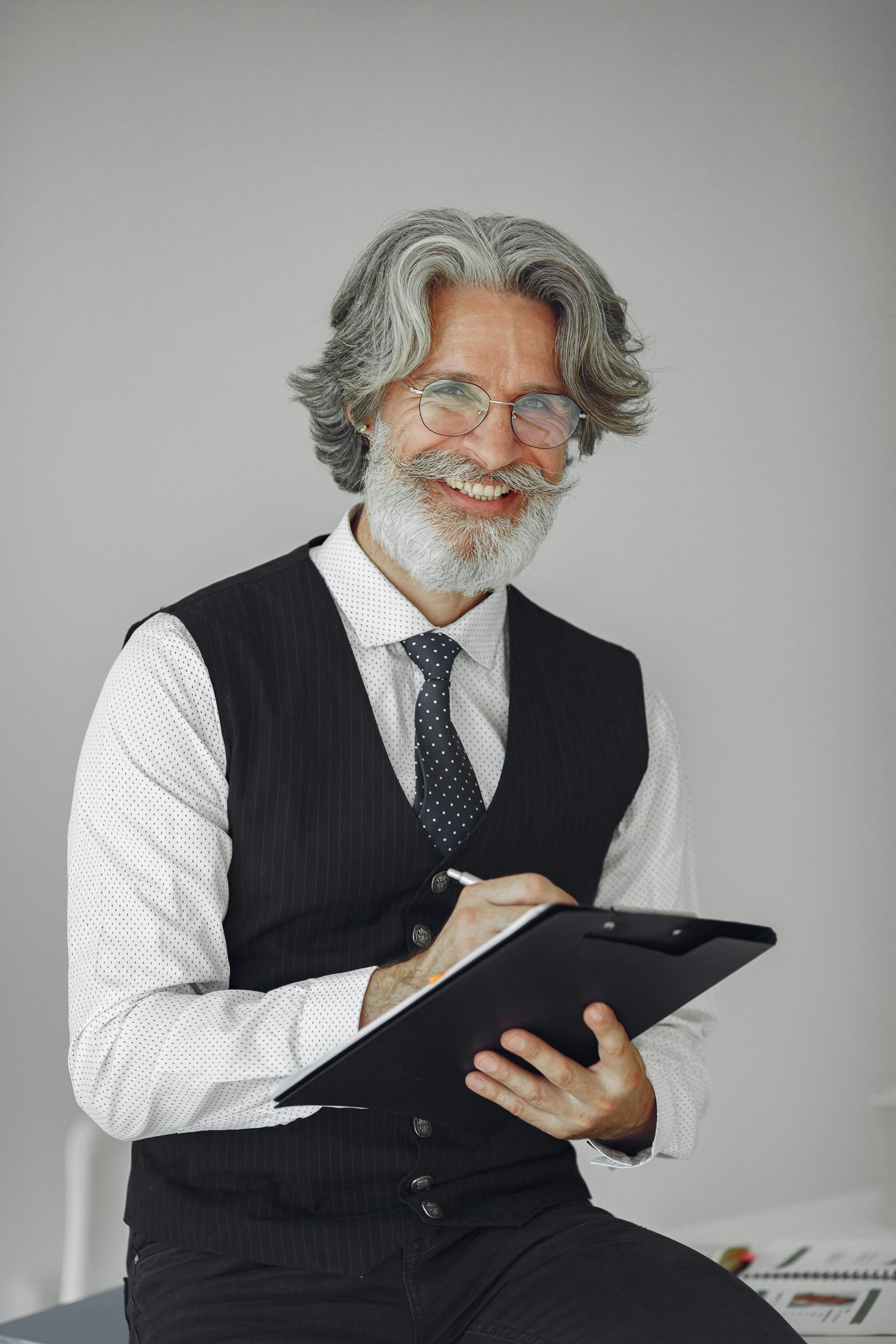 Smiling man with gray hair and beard, wearing glasses, vest, and tie, holding clipboard and pen.